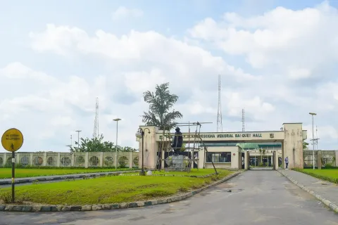 A paved driveway leading to the gated entrance of the Chief D.S.P. Alamieyeseigha Memorial Banquet Hall, with lawns, a palm tree and a cloudy sky