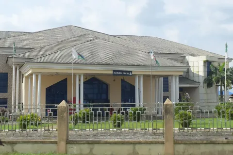 A single-storey hall with a columned entrance and a grey roof behind a fence, under a partly cloudy sky