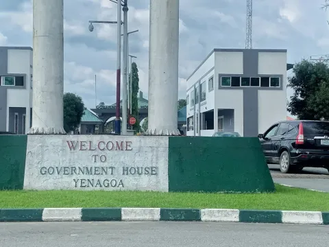 A stone sign reading Welcome to Government House Yenagoa between two tall white pillars beside a road, with a car and buildings behind