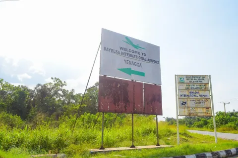 A roadside board reading Welcome to Bayelsa International Airport, Yenagoa, with a smaller notice board and green grass around it