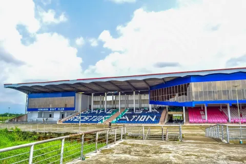 A roofed grandstand with blue signage reading Yenagoa Boat Club and rows of blue, green and pink seats, under a bright sky