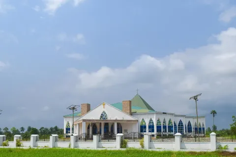 A white building with a green pyramid roof and rows of arched blue windows behind a low wall, with solar street lights and a lawn in front