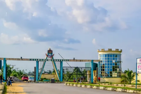 The entrance arch of Niger Delta University with a gatehouse tower, a road, a few vehicles and people near the gate