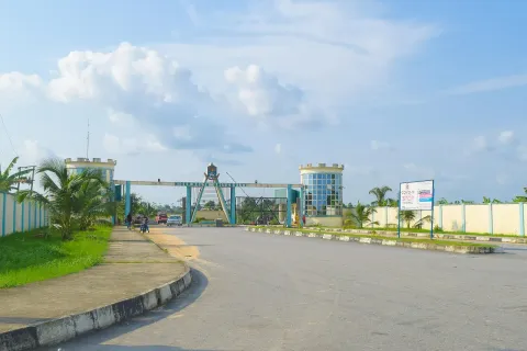 A wide empty road leading to the Niger Delta University main gate, with a castle-style tower and a billboard beside it under a blue sky