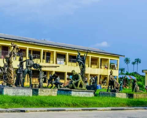 A group of dark statues of figures on a lawn in front of a two-storey yellow building with a palm tree behind