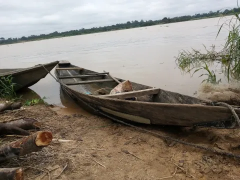 A wooden canoe pulled up on a muddy riverbank with a wide river and an overcast sky behind