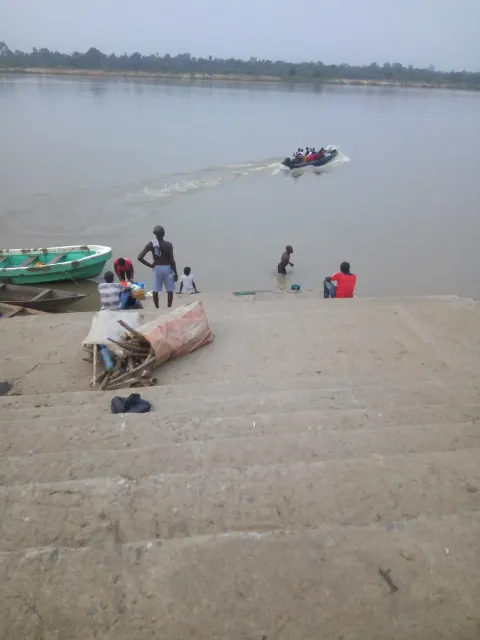 A sandy river landing with a small motorboat crossing hazy water in the distance and a few distant figures on the shore