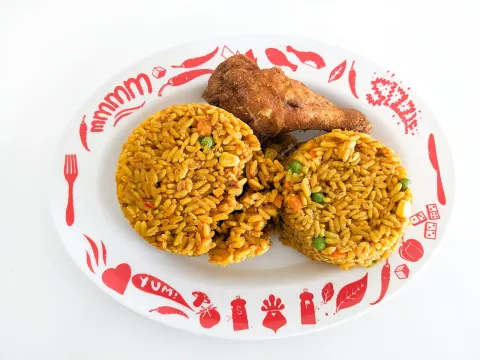 A plate of orange jollof rice with green peas shaped into two mounds beside a fried chicken drumstick