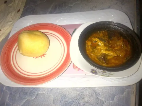 A ball of yellow eba on a plate beside a dark clay pot of orange-yellow banga soup with pieces of fish