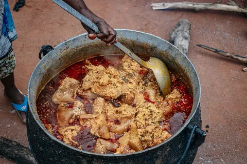A hand holding a ladle over a large black pot of thick red-orange egusi soup with pieces of meat