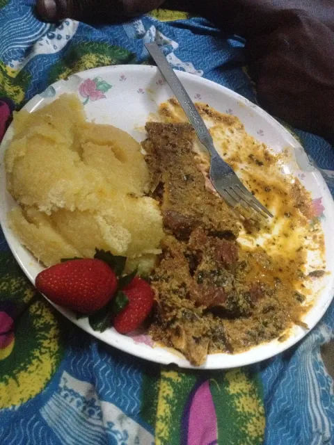 A paper plate with a mound of yellow swallow and egusi soup, a fork and two strawberries, on a colourful patterned cloth