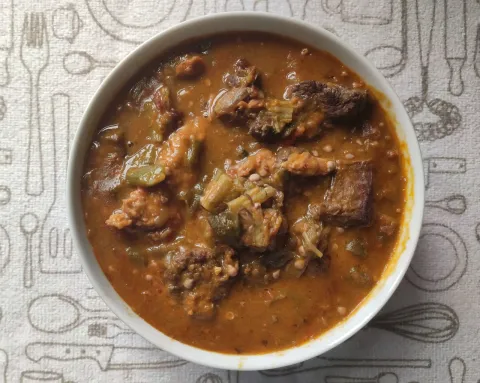 A white bowl of thick brown okro soup with pieces of meat on a patterned paper mat