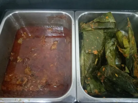 Two steel serving trays, one with red stew and meat and one with moi moi wrapped in green leaves