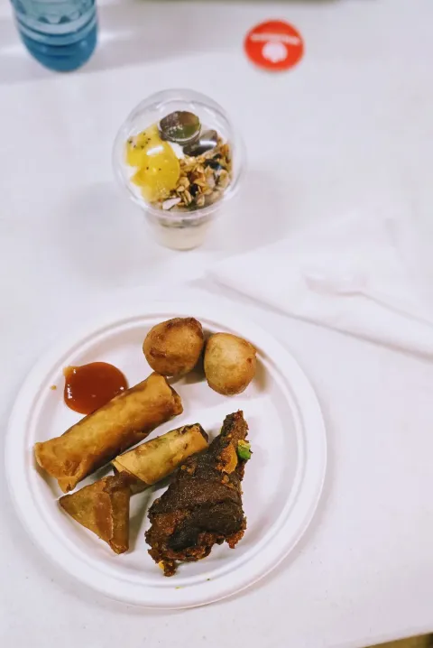 A paper plate of small chops including a spring roll, fried dough balls and spicy chicken, with a cup of yoghurt parfait above it