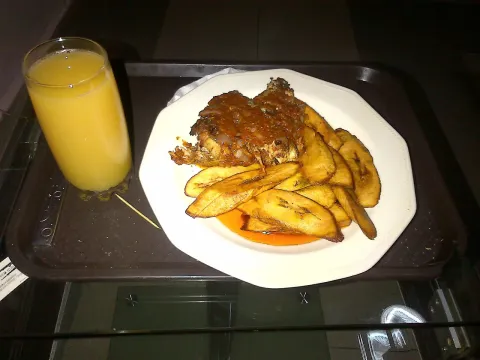 Wedges of fried plantain and a piece of fish in sauce on a white plate, with a glass of orange juice on a tray