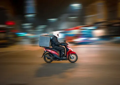 A rider in a white helmet on a red scooter with a grey delivery box passes blurred night-time city lights