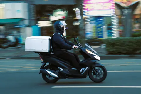 A rider in a helmet and face mask on a black scooter with a white delivery box on the back, riding down a city street