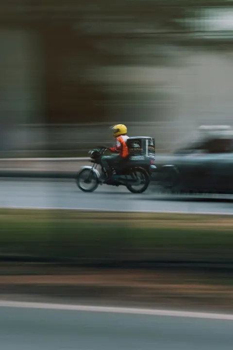 A distant motorcycle rider in an orange jacket carrying a delivery box, blurred by camera motion on a street