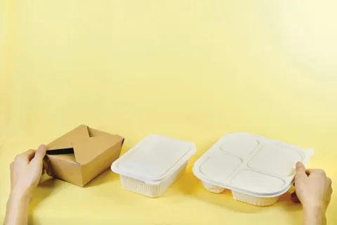 Two hands opening a brown takeaway box beside two white plastic food containers on a yellow background