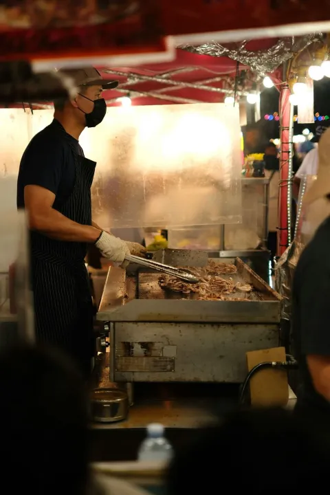 A cook in an apron and face mask grills meat at a lit night-market stall under a red canopy