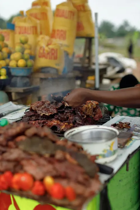 Slices of meat sizzling on an outdoor grill at a roadside stall with tomatoes and fruit stacked beside it